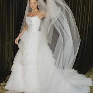 May include: A white strapless wedding dress featuring a structured bodice and a voluminous, multi-layered tulle skirt with tiered ruffles. The gown is paired with a long, sheer white tulle veil that extends outward, creating a flowing effect.
