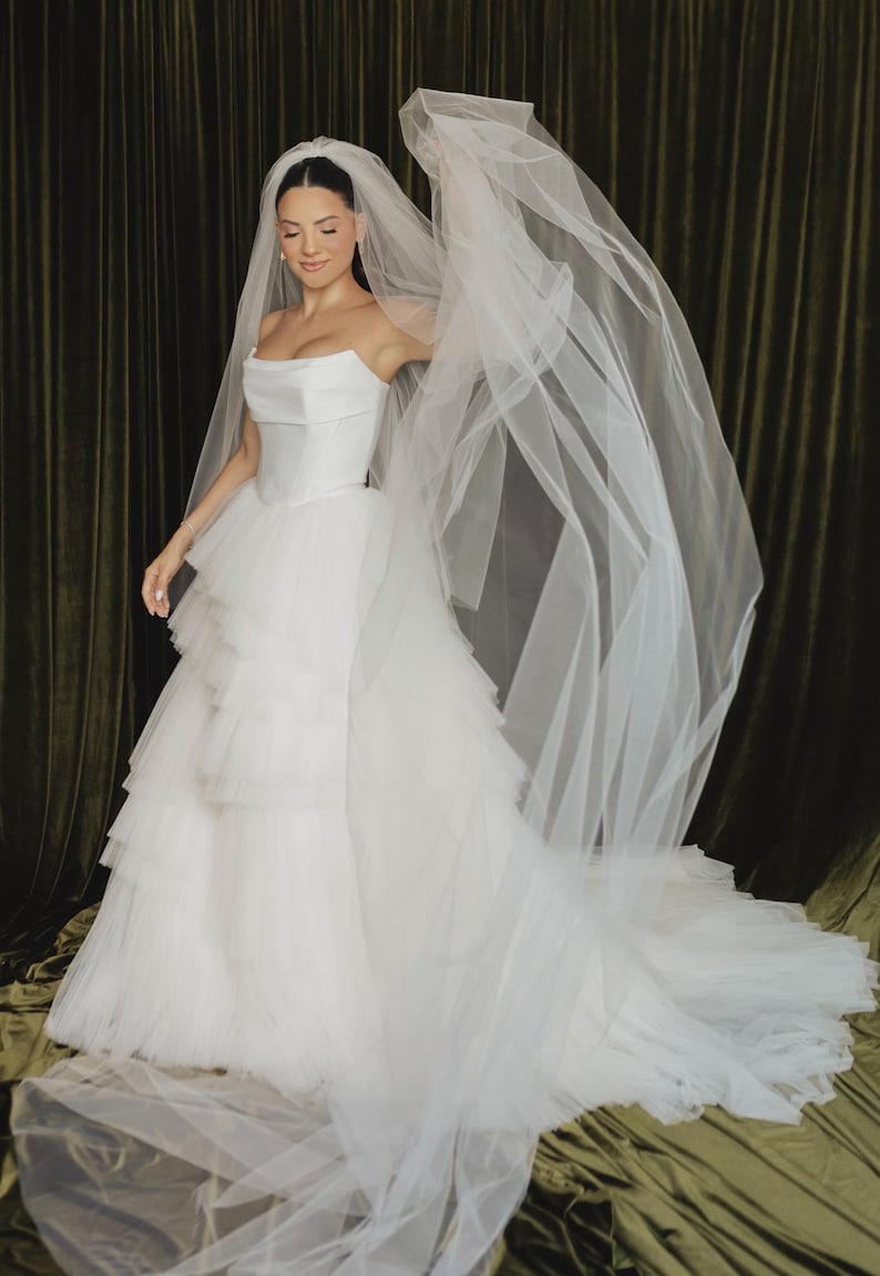 May include: A white strapless wedding dress featuring a structured bodice and a voluminous, multi-layered tulle skirt with tiered ruffles. The gown is paired with a long, sheer white tulle veil that extends outward, creating a flowing effect.