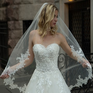 May include: A strapless white wedding dress with intricate floral lace detailing and a flowing tulle skirt. The dress features a sweetheart neckline and is adorned with sequins. A sheer veil with matching lace trim drapes over the shoulders.