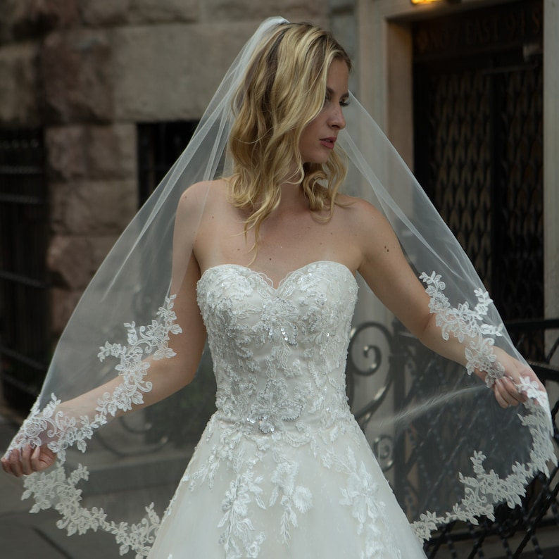 May include: A strapless white wedding dress with intricate floral lace detailing and a flowing tulle skirt. The dress features a sweetheart neckline and is adorned with sequins. A sheer veil with matching lace trim drapes over the shoulders.