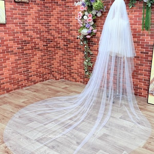May include: A long, white, sheer tulle wedding veil draped over a mannequin. The veil features multiple layers of soft, translucent fabric that extend to the floor, creating a wide, circular train that spreads out across the floor surface.
