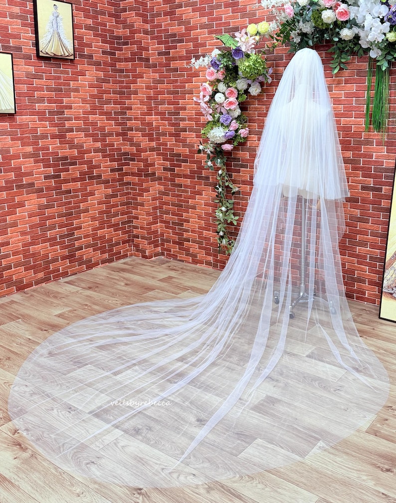 May include: A long, white, sheer tulle wedding veil draped over a mannequin. The veil features multiple layers of soft, translucent fabric that extend to the floor, creating a wide, circular train that spreads out across the floor surface.