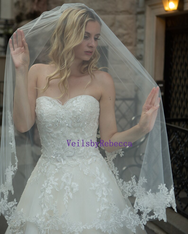 May include: A bride wearing a white strapless wedding dress and a sheer veil. The veil has a delicate lace trim. The dress is adorned with intricate floral embroidery and sequins. The text "VeilsbyRebecca" is visible on the dress.