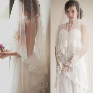 May include: A sheer white bridal veil featuring a scalloped lace trim along the edges. The veil is made of fine tulle fabric and drapes over the head and shoulders, extending down to the waist, showcasing intricate floral lace detailing.