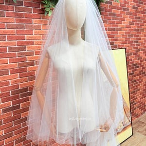 May include: A white, multi-layered tulle wedding veil draped over a mannequin. The sheer fabric features a soft, flowing texture with multiple tiers cascading down the sides. The text "veilsbyrebecca" is printed in small white letters on the veil.