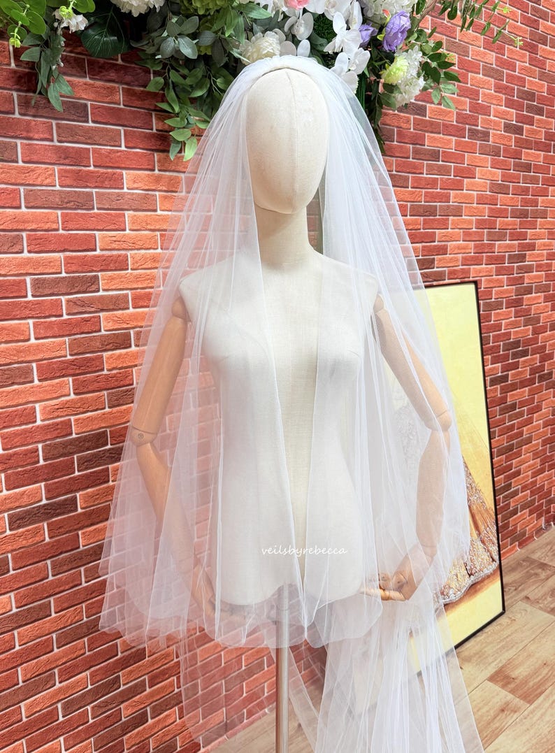 May include: A white, multi-layered tulle wedding veil draped over a mannequin. The sheer fabric features a soft, flowing texture with multiple tiers cascading down the sides. The text "veilsbyrebecca" is printed in small white letters on the veil.