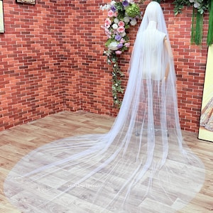 May include: A long, sheer white bridal veil draped over a mannequin. The veil features a soft, flowing tulle fabric that extends onto the floor in a circular train. The design is simple and transparent, showcasing the delicate, lightweight material.