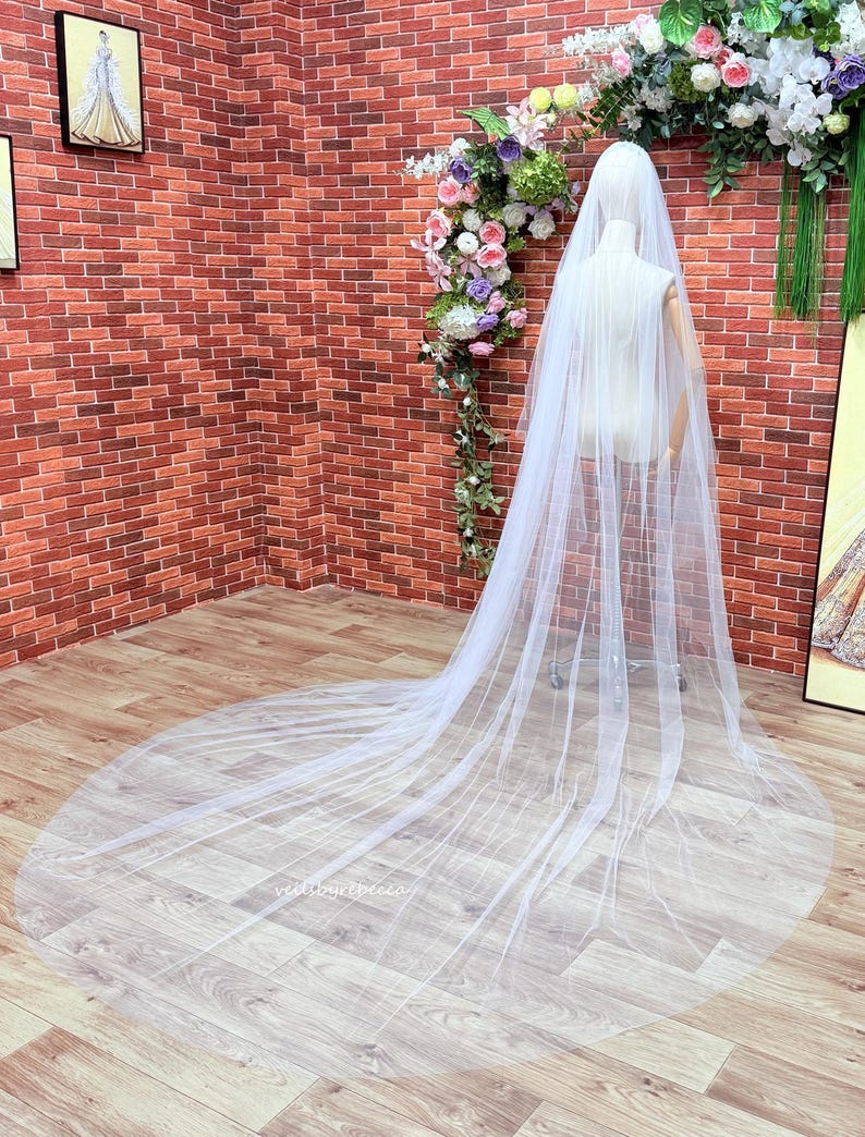 May include: A long, sheer white bridal veil draped over a mannequin. The veil features a soft, flowing tulle fabric that extends onto the floor in a circular train. The design is simple and transparent, showcasing the delicate, lightweight material.