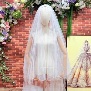 Puede incluir: Un velo de novia blanco y transparente drapeado sobre un maniquí, hecho de una tela de tul suave y superpuesta que cae con gracia desde la cabeza. El velo es liso, sin adornos y translúcido, con el texto "veilsbyrebecca" impreso en pequeñas letras blancas.