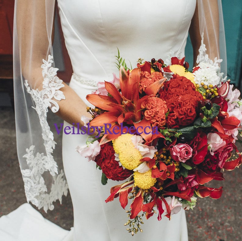 May include: A bride in a white dress holds a vibrant bouquet of red, orange, yellow, and pink flowers. A sheer white veil with delicate lace trim adorns the bride's arm. The text "VeilsbyRebecca" is visible.