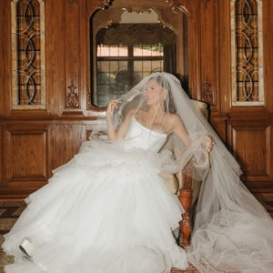 May include: A white strapless wedding dress featuring a fitted bodice and a voluminous, multi-layered tulle skirt. The garment is paired with a long, sheer, floor-length tulle veil that drapes over the head and extends across the floor.