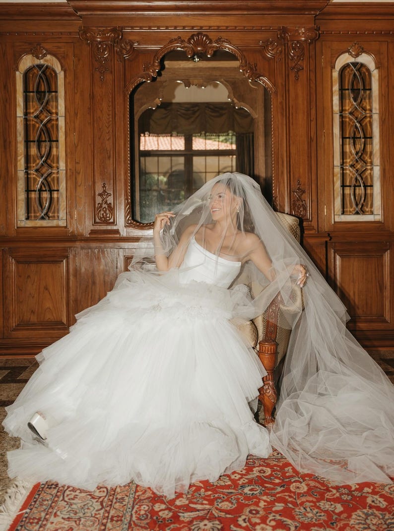 May include: A white strapless wedding dress featuring a fitted bodice and a voluminous, multi-layered tulle skirt. The garment is paired with a long, sheer, floor-length tulle veil that drapes over the head and extends across the floor.