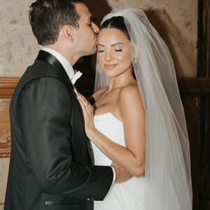 May include: A black tuxedo jacket with a white shirt and bow tie worn by one individual, and a strapless white wedding dress with a sheer tulle veil worn by another individual. The individual in the tuxedo kisses the forehead of the other.