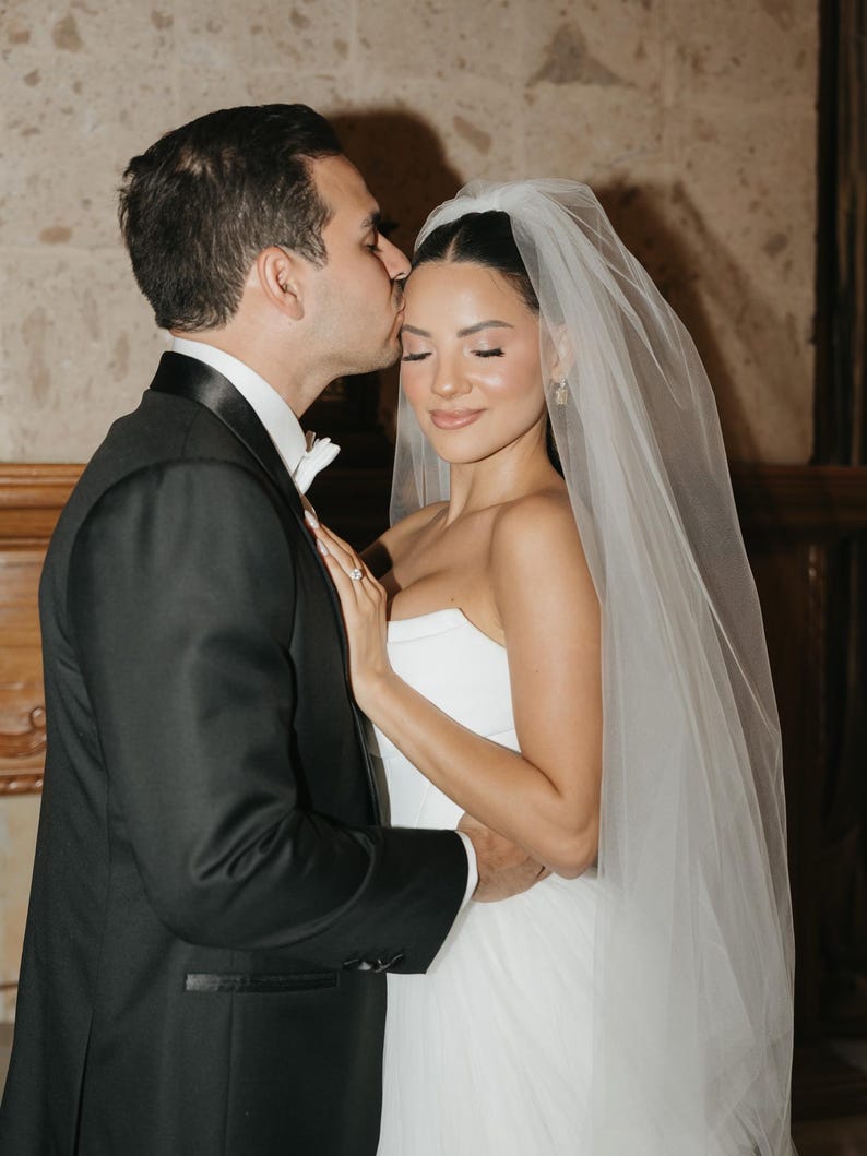 May include: A black tuxedo jacket with a white shirt and bow tie worn by one individual, and a strapless white wedding dress with a sheer tulle veil worn by another individual. The individual in the tuxedo kisses the forehead of the other.