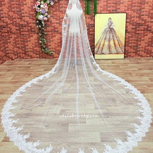 May include: A long, white bridal veil with a delicate lace trim. The veil is displayed on a mannequin, showcasing its length and intricate design. The words "veilsbyrebecca" are visible on the veil. A framed illustration of a bride is in the background.