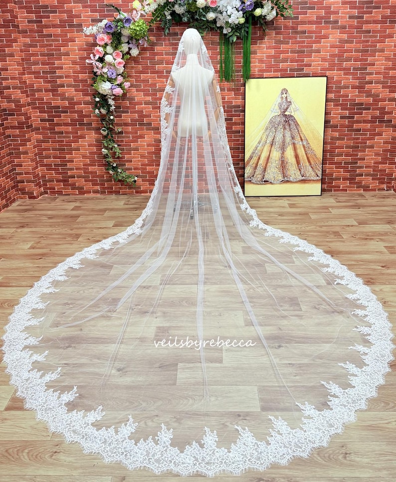 May include: A long, white bridal veil with a delicate lace trim. The veil is displayed on a mannequin, showcasing its length and intricate design. The words "veilsbyrebecca" are visible on the veil. A framed illustration of a bride is in the background.