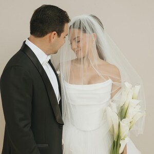 May include: A black tuxedo with a white shirt and a white bow tie worn next to a white strapless gown with a layered tulle skirt and a sheer veil, accompanied by a bouquet of white calla lilies with green stems wrapped in a white ribbon.