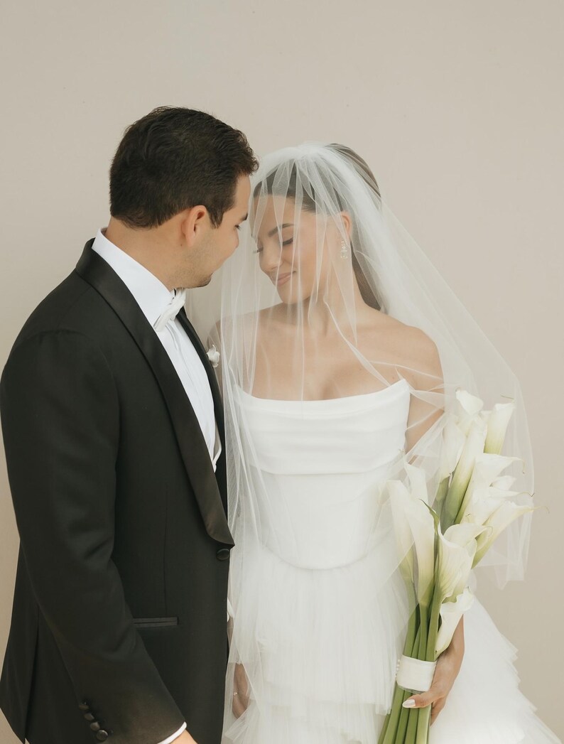 May include: A black tuxedo with a white shirt and a white bow tie worn next to a white strapless gown with a layered tulle skirt and a sheer veil, accompanied by a bouquet of white calla lilies with green stems wrapped in a white ribbon.