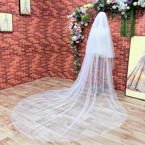 May include: A long, sheer white bridal veil made of fine tulle fabric. The veil features a multi-layered design that cascades down from a central support, spreading out across the floor in a wide, circular train. The material is translucent.