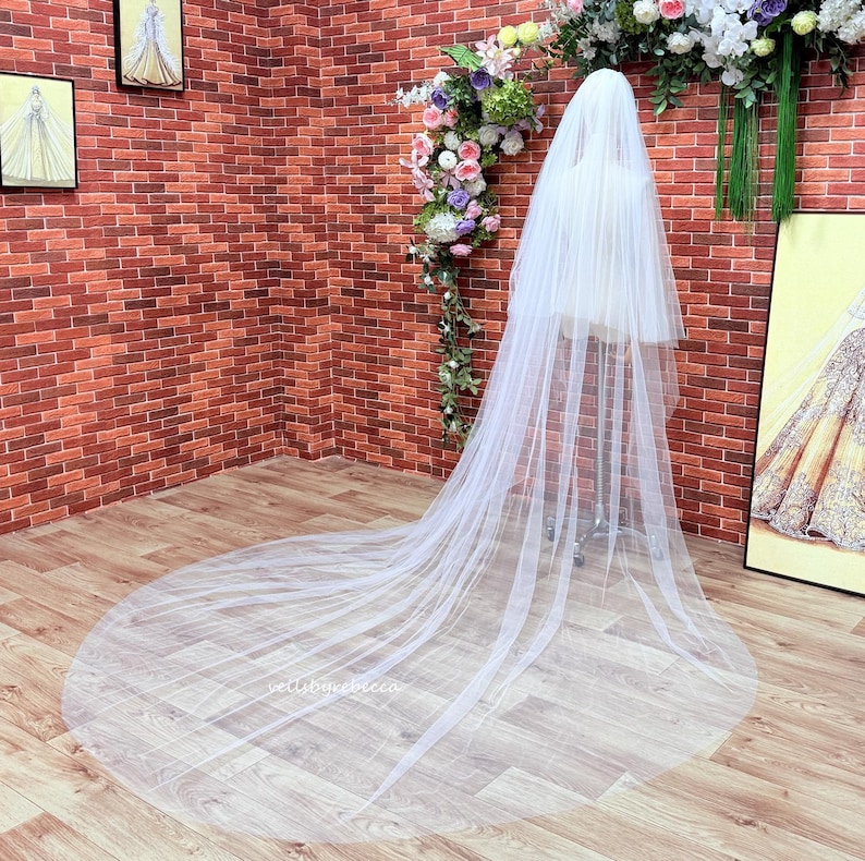 May include: A long, sheer white bridal veil made of fine tulle fabric. The veil features a multi-layered design that cascades down from a central support, spreading out across the floor in a wide, circular train. The material is translucent.