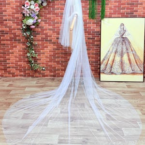 May include: A long, sheer white bridal veil made of fine tulle fabric. The veil features multiple layers that cascade down from the head, creating a dramatic floor-length train. The text "veilsbyrebecca" is printed in small white letters on the fabric.