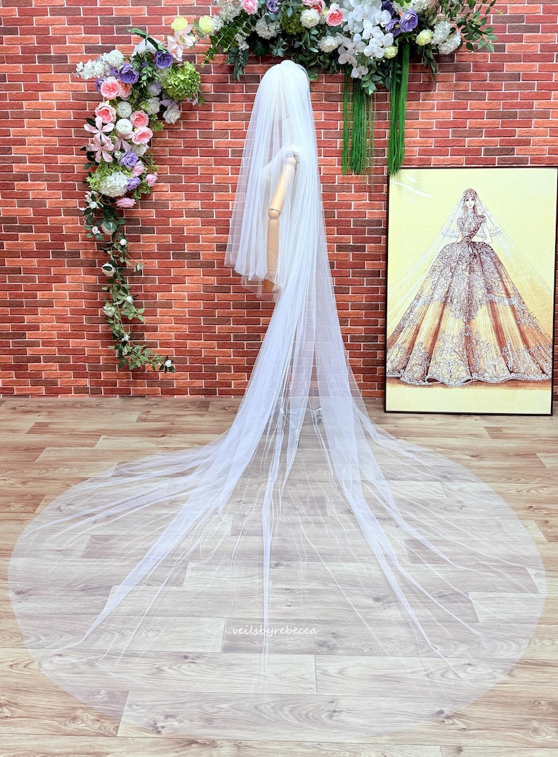 May include: A long, sheer white bridal veil made of fine tulle fabric. The veil features multiple layers that cascade down from the head, creating a dramatic floor-length train. The text "veilsbyrebecca" is printed in small white letters on the fabric.