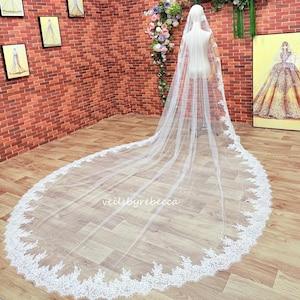 May include: A long, white bridal veil with a delicate lace trim. The veil is displayed on a mannequin, showcasing its length and intricate design. The text "veilsbyrebecca" is visible on the veil.