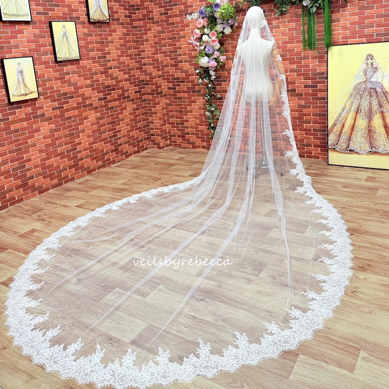 May include: A long, white bridal veil with a delicate lace trim. The veil is displayed on a mannequin, showcasing its length and intricate design. The text "veilsbyrebecca" is visible on the veil.