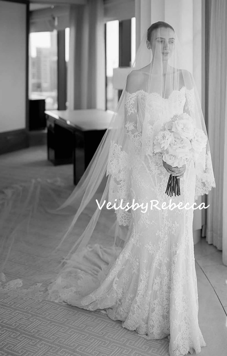 May include: A bride wearing a white lace wedding dress with a long veil and holding a bouquet of white flowers. The text "Veils by Rebecca" is visible in the image.