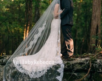 May include: A bride wearing a white wedding dress with a long, flowing veil with lace trim. The veil is draped over the bride's head and shoulders, and the lace trim is visible along the edges of the veil. The bride is standing next to a groom in a dark suit. The couple is standing on a log in a forest setting.
