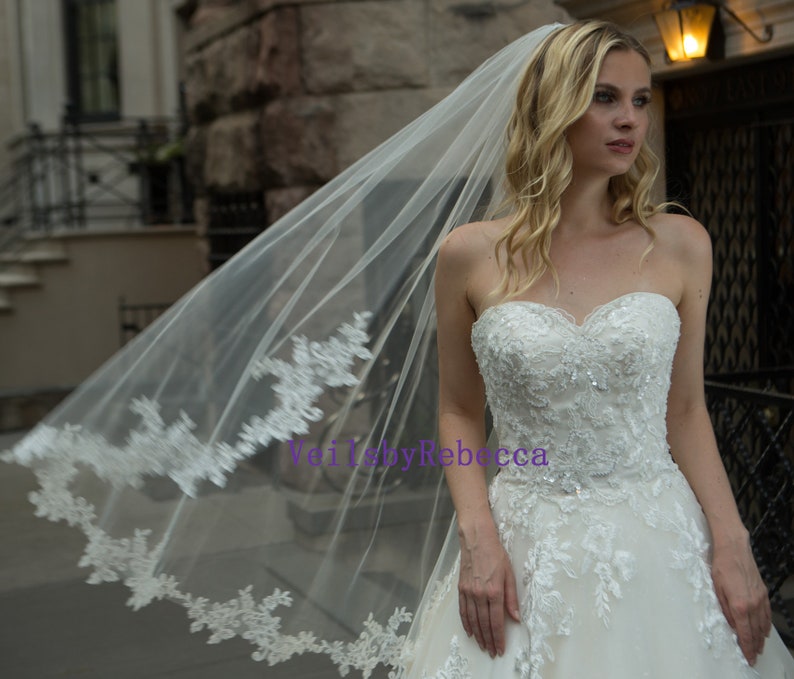 May include: A strapless white wedding dress with intricate lace and sequin detailing. A long, sheer veil with delicate lace trim flows in the wind. The text "Veil by Rebecca" is visible.