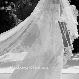 May include: A white bridal veil with delicate lace trim. The veil is flowing in the wind, creating a romantic and ethereal look. The text "Veils by Rebecca" is visible in the lower left corner of the image.