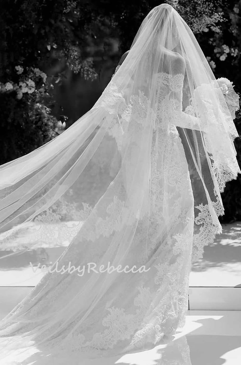 May include: A white bridal veil with delicate lace trim. The veil is flowing in the wind, creating a romantic and ethereal look. The text "Veils by Rebecca" is visible in the lower left corner of the image.