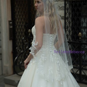 May include: A strapless ivory wedding dress with lace detailing and a long, sheer veil. The veil is adorned with a sparkling headpiece. The dress features a corset back and a full skirt, creating an elegant bridal look.
