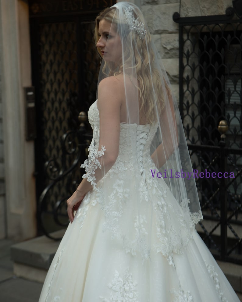 May include: A strapless ivory wedding dress with lace detailing and a long, sheer veil. The veil is adorned with a sparkling headpiece. The dress features a corset back and a full skirt, creating an elegant bridal look.