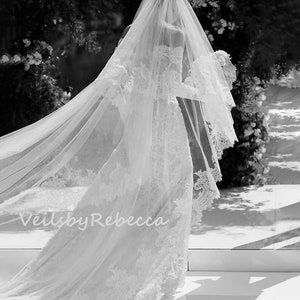 May include: A white wedding veil with lace trim and a long train. The veil is flowing in the wind. The text "Veils by Rebecca" is visible in the bottom left corner of the image.