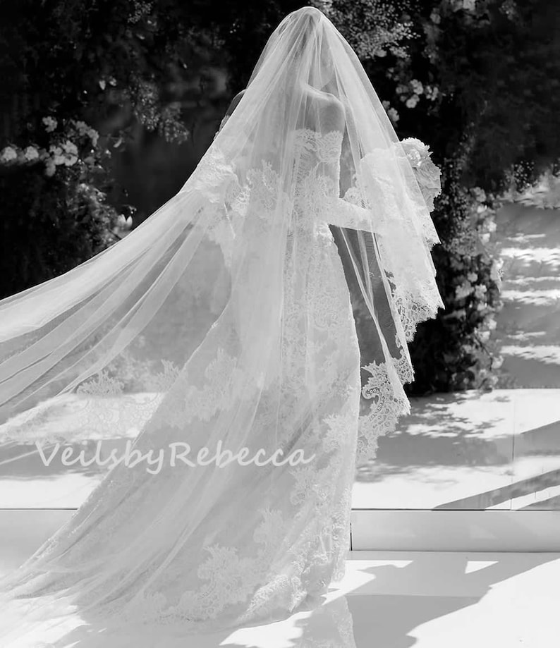 May include: A white wedding veil with lace trim and a long train. The veil is flowing in the wind. The text "Veils by Rebecca" is visible in the bottom left corner of the image.