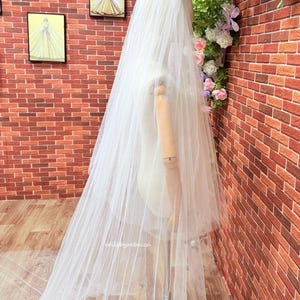 May include: A long, sheer white wedding veil draped over a wooden mannequin. The veil is made of fine, translucent tulle fabric that extends to the floor. The text "veilsbyrebecca" is printed in small, white, lowercase letters on the fabric.