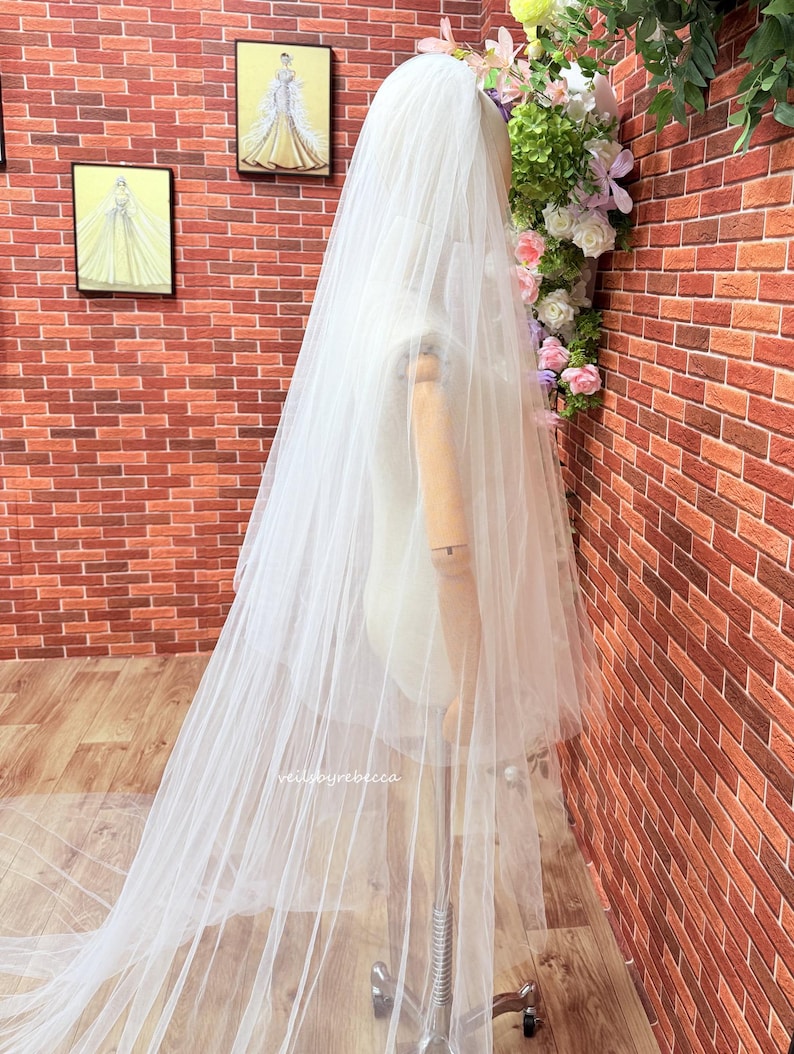 May include: A long, sheer white wedding veil draped over a wooden mannequin. The veil is made of fine, translucent tulle fabric that extends to the floor. The text "veilsbyrebecca" is printed in small, white, lowercase letters on the fabric.