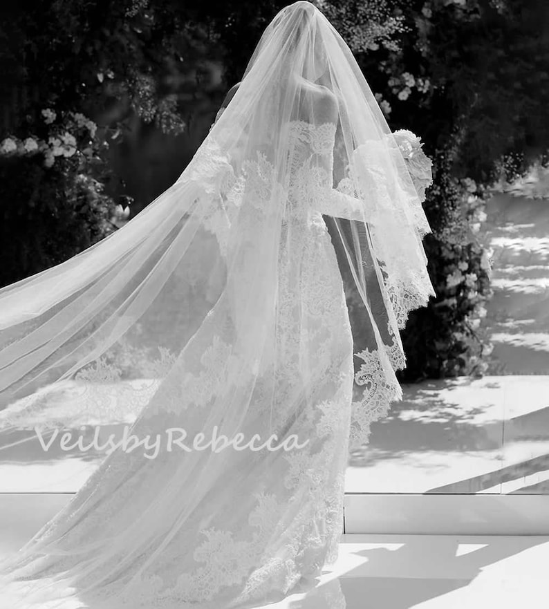 May include: A bride wearing a white wedding dress with a long, flowing veil. The veil is made of delicate lace and has a scalloped edge. The bride is walking down the aisle, and her veil is flowing behind her. The text "Veils by Rebecca" is visible in the bottom left corner of the image.