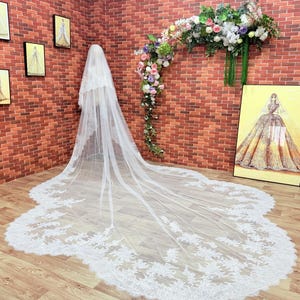 Puede incluir: Un velo de novia blanco largo con intrincados detalles de encaje floral en los bordes. El velo se exhibe en un maniquí, mostrando su diseño fluido. El fondo presenta una pared de ladrillo, arreglos florales y obras de arte enmarcadas.