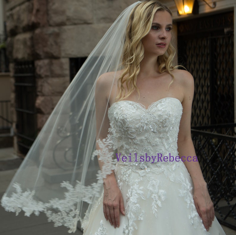 May include: A white bridal veil with delicate lace trim and a strapless wedding dress with intricate beadwork. The veil is long and flowing, creating an elegant and romantic look for a wedding.