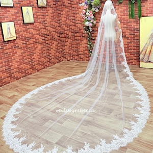 May include: A long, white bridal veil with detailed lace trim. The veil is draped on a mannequin, highlighting its length and delicate design. The text "veilbyrebecca" is visible on the veil.