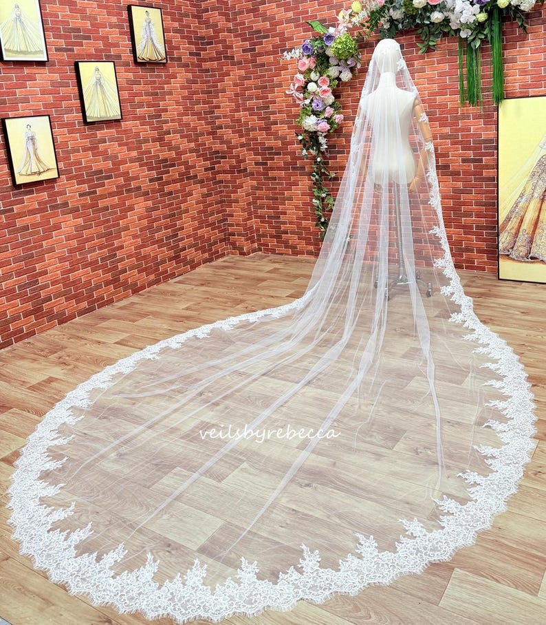 May include: A long, white bridal veil with detailed lace trim. The veil is draped on a mannequin, highlighting its length and delicate design. The text "veilbyrebecca" is visible on the veil.