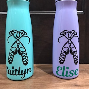 May include: Two personalized water bottles with a teal and purple finish. Each bottle features a black silhouette of Irish dance shoes and a name, "Caitlyn" and "Elise", in black lettering.