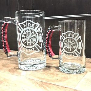 May include: Two glass beer mugs with black and red paracord handles. Both mugs feature an etched design of a fire department logo with the words "FIRE DEPT."