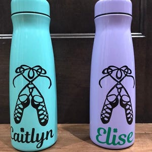May include: Two cylindrical water bottles with matching caps. The left bottle is light blue with a black graphic of ballet slippers and the name Caitlyn in black script. The right bottle is light purple with a black graphic of ballet slippers and the name Elise in green script.
