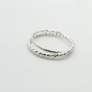 May include: A silver ring with a twisted rope design on one band and a smooth band above it. The ring is set against a white background, highlighting its metallic shine. This piece of jewelry is a simple yet elegant accessory.