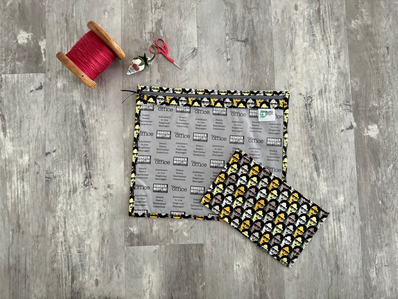 Pode incluir: Uma bolsa transparente com fecho de correr de pl&aacute;stico com um forro de tecido preto e amarelo com um padr&atilde;o repetido dos personagens do programa de televis&atilde;o "The Office". A bolsa est&aacute; aberta e uma bolsa de tecido mais pequena de cor preta e amarela com o mesmo padr&atilde;o est&aacute; dentro.