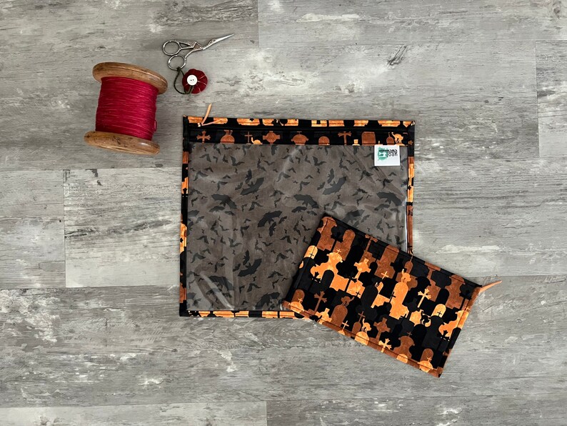 Pode incluir: Duas bolsas com fecho de correr em tecido com tema de Halloween. Uma bolsa &eacute; preta com silhuetas laranja e castanhas de l&aacute;pides e cruzes. A outra bolsa &eacute; preta com silhuetas cinzentas de morcegos. Ambas as bolsas t&ecirc;m janelas de pl&aacute;stico transparente.